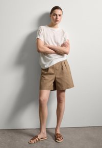 Lässiges Outfit mit einem hellgrauen Leinen-T-Shirt und braunen, gefalteten Shorts, kombiniert mit braunen, sandalenartigen Schuhen. Die Wand ist hellgrau.