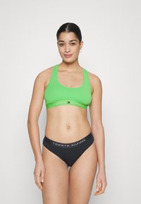Soutien-gorge de sport vert avec de larges bretelles et une bande logo blanche, associé à un bas de bikini noir avec une taille logo. Texture lisse.