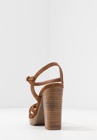 Brown suede high-heeled sandal with wooden block heel and ankle strap, photographed against a white corner background.