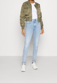 Vihreä satiininen bomber-takki, jossa resorit hihansuissa, yhdistetty vaaleansinisiin kapealahkeisiin farkkuihin ja valkoisiin lenkkareihin; minimalistinen, istuva malli.