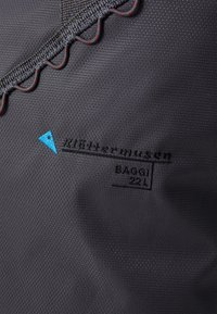 Tissu noir texturé avec des coutures rouges et un détail de logo bleu. Présente un branding en relief : "Kletterergemüse BAGGI 22L".