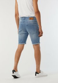 Shorts en denim bleu clair, présentant un ourlet roulé, deux poches arrière et une étiquette de marque au niveau de la taille. Porté avec des baskets blanches.