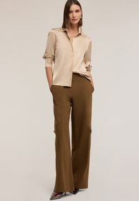 Beżowa bluzka z kolnierzykiem ozdobionym ćwiekami, w zestawieniu z brązowymi spodniami typu cargo o szerokich nogawkach, które mają kieszenie i zamki. Lśniące brązowe obcasy dopełniają stylizację.