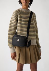 Schwarze Umhängetasche mit goldenem Reißverschluss und Logo, kombiniert mit einem gestreiften beigen und grauen Pullover und einem plissierten khakifarbenen Rock.