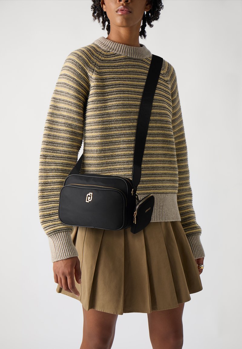 Schwarze Umhängetasche mit goldenem Reißverschluss und Logo, kombiniert mit einem gestreiften beigen und grauen Pullover und einem plissierten khakifarbenen Rock.