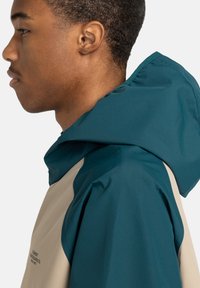 Capuchon jasje met een twee-kleurig ontwerp in teal en beige. Gemaakt van glad materiaal met een strakke, op buitenactiviteiten gerichte uitstraling.