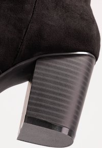 Czarny zamszowy botek wokół kostki z masywnym, żłobkowanym obcasem z gładkim metalowym akcentem u podstawy. Skupienie na fakturze i geometrycznym kształcie.