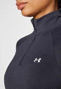 Crna zip-up atletska majica napravljena od glatkog materijala, sa visokim okovratnikom i malim bijelim Under Armour logom na prsima.