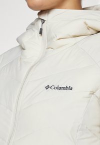 Hvid isoleret jakke med høj krave, lynlås foran og diagonalt quiltet tekstur. Har Columbia-logoet på brystet.
