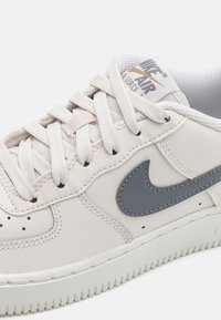 Sneakers Nike Air Force 1 ve světle béžové kůži, s šedým koženým Swooshem, plochými bílými tkaničkami a texturovanou gumovou podrážkou.