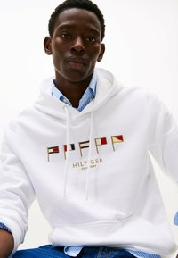 Witte hoodie met geborduurde vlagmotieven in rood, blauw en geel, met daaronder "HILFIGER" en "Since 1985" in goud. Getextureerde katoenstof.