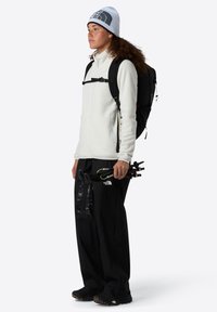 Jachetă albă din fleece, pantaloni negri cu accente impermeabile, rucsac negru și căciulă gri; ținând bețe de trekking.