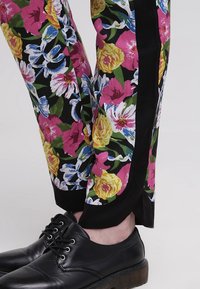 Bloemmotieven broeken tonen levendige roze, gele en blauwe bloemen op een zwarte achtergrond, met een contrasterend zwart zijpaneel. Zwarte schoenen worden gedragen.