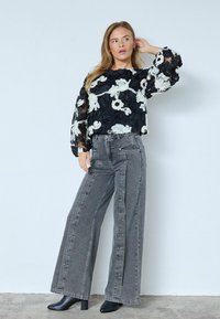 Melna ziedu izšuvuma blūze ar caurspīdīgām piedurknēm, saskaņota ar augsto jostasvietu pelēkiem platām biksēm ar pogu akcentiem un melnām potītēm.