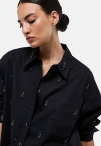 Chemise noire à design oversize, ornée de petites perles noires et de sequins, avec un boutonnage à l'avant et une poche sur la poitrine.