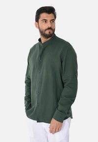 Camicia verde con colletto alla mandarina, maniche lunghe e tessuto leggermente strutturato. I bottoni sono abbinati nel colore. Indossata con pantaloni bianchi.