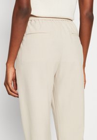 Beige skräddarsydda byxor med elastiskt midjeband. Har två bakfickor, slät tygtextur och raka ben.