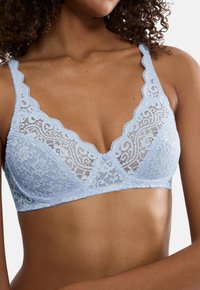 Ženska v svetlo modri čipkasti modrček z cvetličnimi motivi in valovitimi robovi, ki prikazuje zgornji del telesa in kodraste lase.