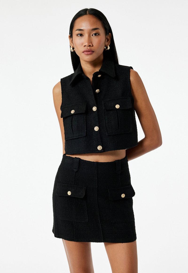 Sort cropped vest og matchende nederdel i struktureret stof. Har guldknapper og praktiske lommer. Struktureret design med krave.