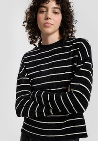Czarnobiały sweter w paski z okrągłym dekoltem, o fakturowanej tkaninie, luźnym kroju i długich rękawach. Brak widocznych wzorów lub akcentów.