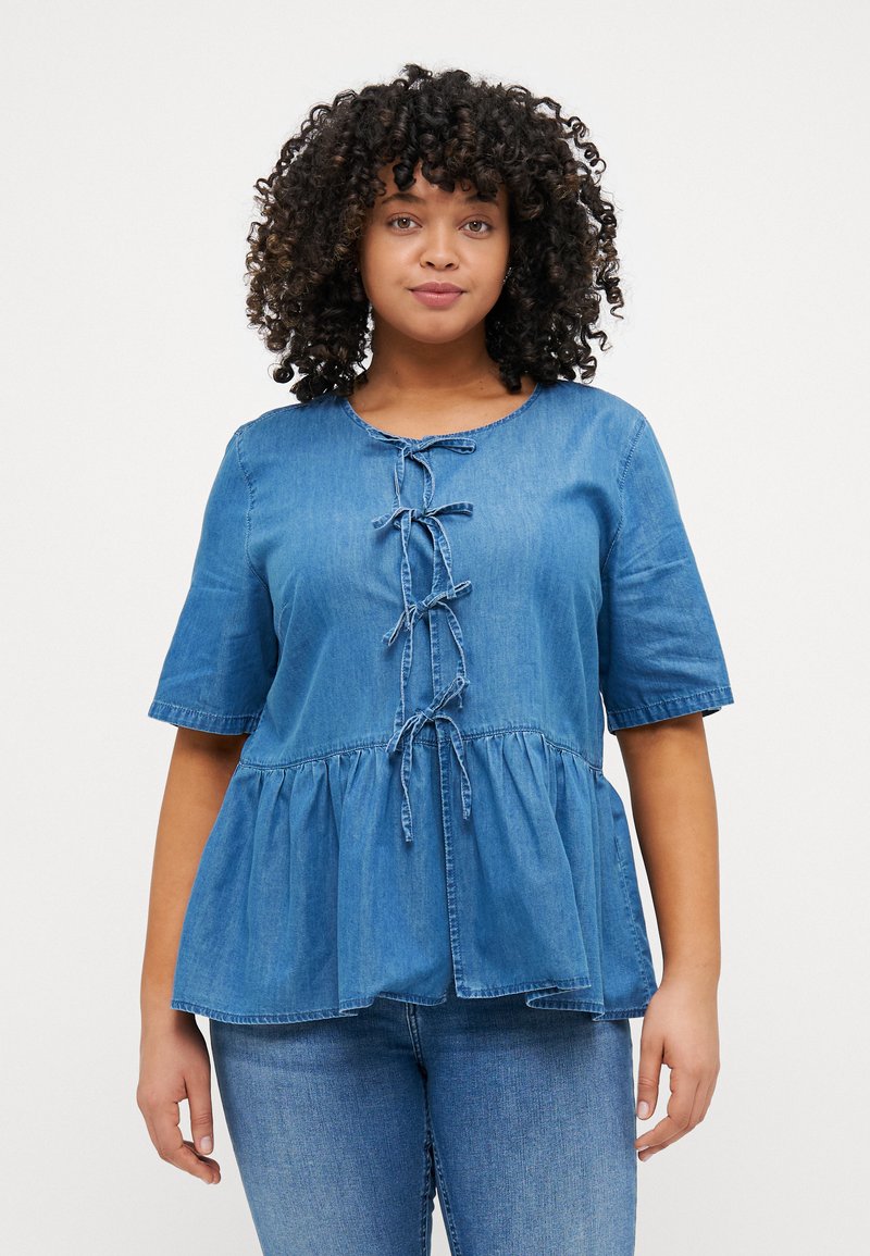 Top peplum de mezclilla azul claro con escote redondeado, mangas cortas y detalle de amarre en el frontal. Textura suave, corte informal y costuras acentuadas.