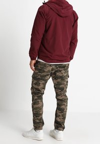 Bordowa lekką kurtka z kapturem, połączona z zielonymi spodniami cargo w kamuflażu i białymi sneakersami. Widoczne detale kieszeni i zwężane wykończenia nogawek.