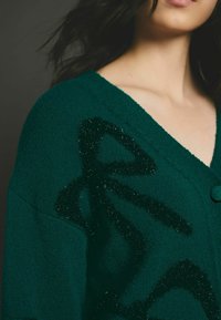 Gilet vert avec une finition texturée, présentant un grand motif noir duveteux. Il a un décolleté en V et un accent de bouton.