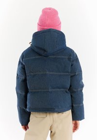 Persoon in een donkerblauwe gewatteerde jas met capuchon, beige broek en een felroze gebreide muts, met de rug naar de camera op een witte achtergrond.