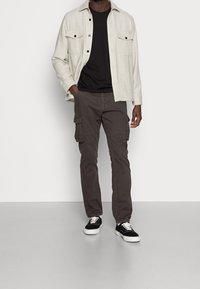 Ljusgrå skjorta med knappar över en svart t-shirt, tillsammans med mörka cargobyxor och svarta sneakers med vita sulor. Enkelt, avslappnat utseende.