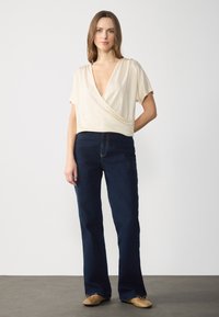 Haut enroulé beige à manches courtes et décolleté en V profond, associé à un jean flare bleu foncé et des chaussures beiges. Design simple et texture douce.