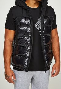 Mężczyzna w czarnej, błyszczącej kamizelce puchowej nosi czarną koszulkę z grafiką, w połączeniu z jasnoszarymi spodniami dresowymi, odsłaniając tatuaż na lewej ręce.