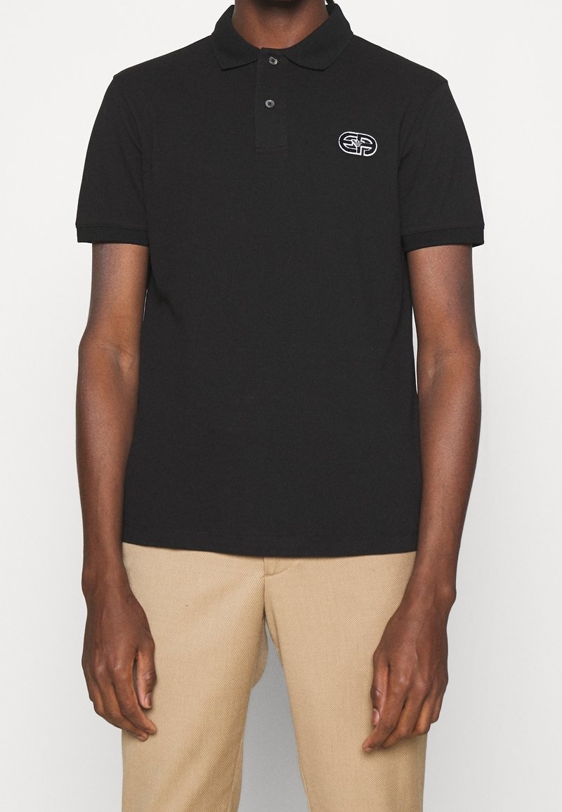 Personne portant un polo noir à manches courtes avec un petit logo blanc sur la poitrine gauche et un pantalon beige.