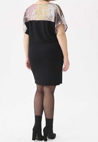 Žena v černých šatech po kolena se zdobeným topem a odhalenými rameny, průhlednými černými punčochami a černými vysokými kotníkovými botami, otočená zády.