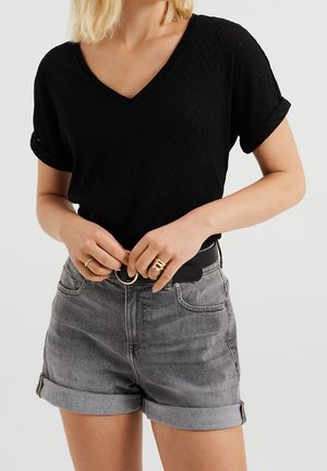 Kvinde iført en sort tekstureret V-hals T-shirt og opdagede grå denimshorts, der spænder et sort bælte med gyldne ringe.