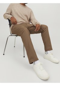 Maglione beige chiaro, pantaloni marroni a gamba dritta e scarpe da ginnastica bianche indossati da una persona seduta su una sedia. Sfondo neutro.
