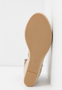 Semelle d’une chaussure compensée beige avec dessous en caoutchouc texturé, appuyée contre un mur blanc d’angle, montrant la pointure 4.