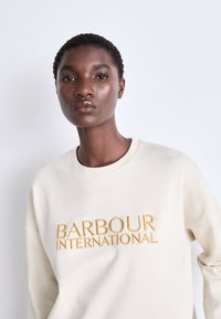 Tejfehér pulóver, melyen arany hímezéssel szerepel a "BARBOUR INTERNATIONAL" felirat, kerek nyakkivágással, laza szabással és hosszú ujjakkal.