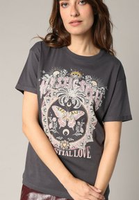 Camiseta de algodón gris con un gráfico circular de una mariposa con acentos florales y el texto "MYSTIC SPEEL" y "CESTIAL LOVE" en rosa.