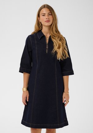 Femme aux longs cheveux ondulés portant une robe en jean foncé, longueur genou, avec une fermeture éclair devant et des manches jusqu’aux coudes, accessoirisée de bijoux dorés.