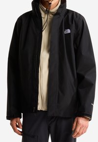 Personne portant une veste North Face noire zippée par-dessus une polaire beige zippée, les mains détendues le long du corps sur un fond blanc.
