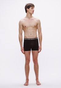 Μαύρα βαμβακερά ανδρικά εσώρουχα boxer με ελαστική μέση. Το μοντέλο στέκεται όρθιο, παρουσιάζοντας τη εφαρμογή και τη δομή, πάνω σε λευκό απλό φόντο.