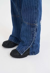 Niebieskie dżinsy z pionowymi paskami, z postrzępionym brzegiem i zatrzaskami przy mankietach. W połączeniu z czarnymi butami wsuwanymi.