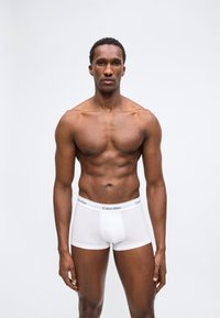 Άνδρας μοντέλο με μυώδη κορμί που φοράει λευκά μπόξερ Calvin Klein, στέκεται μπροστά σε απλό ανοιχτό γκρι φόντο.