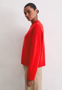 Czerwony krótki sweter z prążkowanym dołem i mankietami, wykonany z miękkiego materiału. Model stoi bokiem na neutralnym tle.