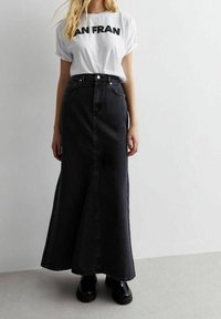 Femme portant un t-shirt blanc "SAN FRAN" rentré dans une jupe maxi en denim noir taille haute avec des chaussures à plateforme noires sur un sol gris.