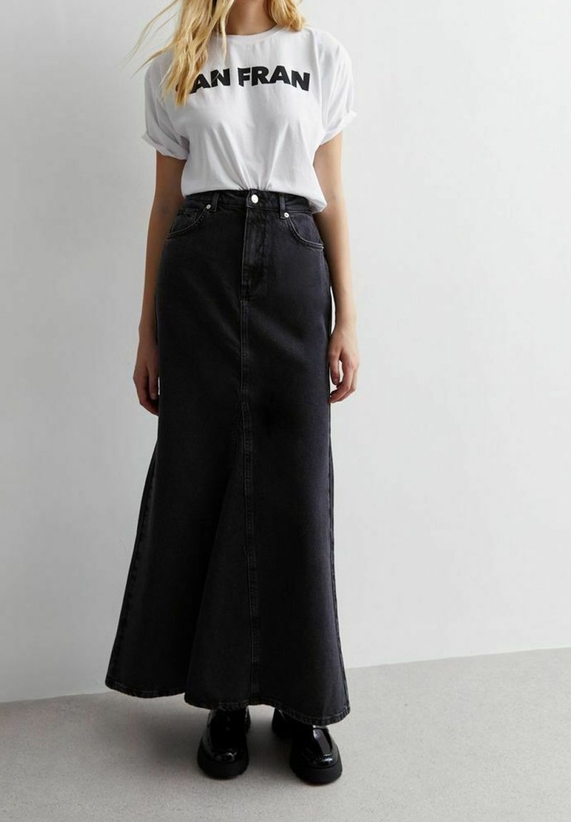 Femme portant un t-shirt blanc "SAN FRAN" rentré dans une jupe maxi en denim noir taille haute avec des chaussures à plateforme noires sur un sol gris.