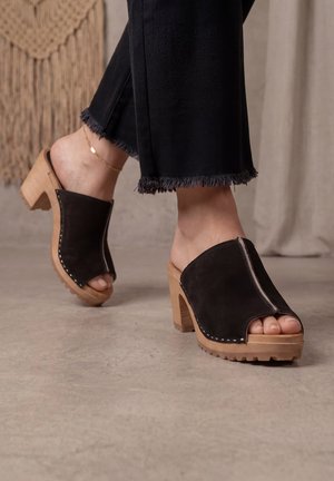 Pieds chaussés de mules ouvertes en daim noir avec des talons hauts en bois et un pantalon noir aux bords effilochés, debout sur un sol lisse.