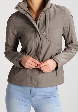 Person fastening zipper on fitted light brown jacket with multiple pockets, wearing light blue jeans against plain background.