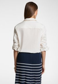 Camicia bianca a maniche lunghe con polsini arrotolati, abbinata a una gonna plissettata blu navy con strisce orizzontali bianche. Tessuto liscio, design con colletto.