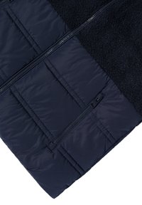 Chaqueta azul marino con un diseño acolchado y un panel superior de forro polar suave, cierre de cremallera completa y bolsillo lateral con cremallera. Superficie texturizada en todo el material.